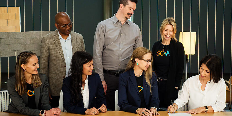 A group of people look at a woman who is signing a contract at a table.