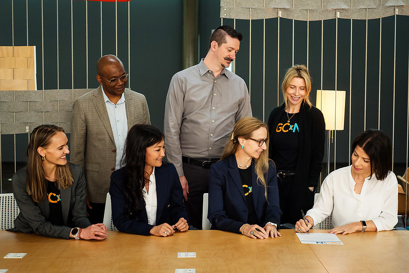 Why the New GCxN Cohort Is Just the Start A group of people look at a woman who is signing a contract at a table.