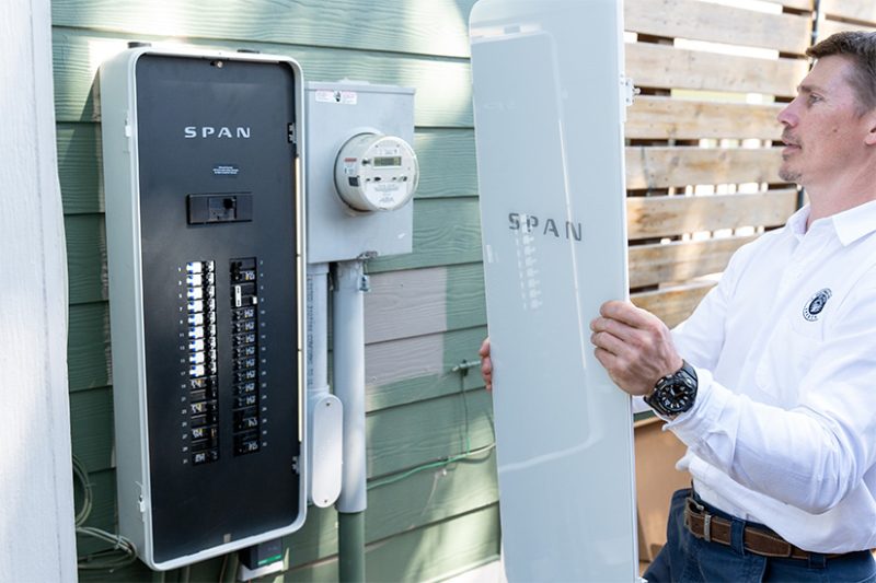 SPAN A man holds the cover of a power panel next to the panel hanging on the outside of a home.
